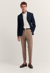 Blazer blu navy, polo bianco e pantaloni beige chiaro. Mocassini marroni e cintura marrone. Design slim fit con pantaloni plissettati. Texture morbide.