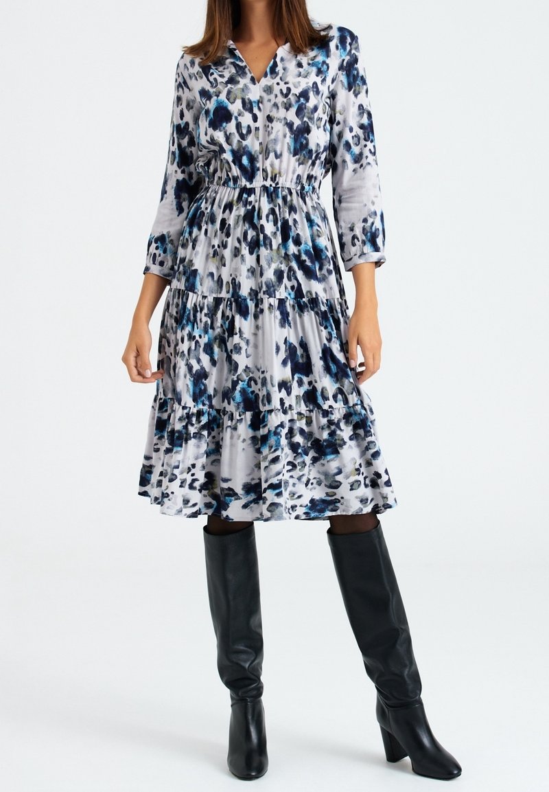 Femme portant une robe midi à pois blancs, bleus et noirs avec des manches trois-quarts et des bottes noires à talons hauts, jusqu'aux genoux.