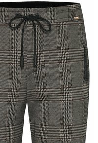 Pantalon à carreaux noirs, beiges et blancs avec une taille à cordon noir et une poche zippée sur le côté droit.