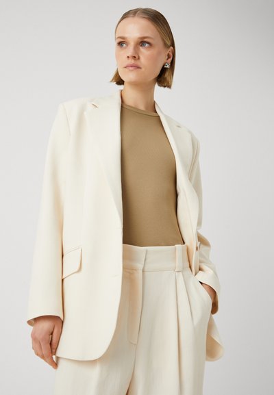 Blazer crema con vestibilità su misura, abbinato a un top beige a costine. L'outfit presenta pantaloni a gamba larga e tasche strutturate.