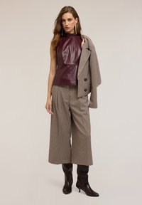 Donna che indossa un top in pelle bordeaux senza maniche, pantaloni a gamba larga a quadri, tiene in spalla un blazer coordinato a quadri e stivali neri con tacco.