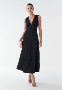 Robe maxi en lin noir avec un décolleté en V profond et une taille cintrée, présentant une finition texturée. Associée à des accessoires dorés et des talons dénudés.