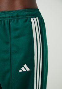 Groene sportshorts van ademend materiaal, met drie witte verticale strepen en een Adidas-logo op de linker bovenbeen.