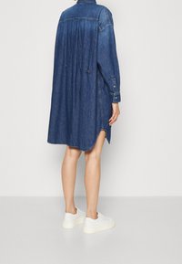 Donkerblauwe denim jurk met lange mouwen, knoopsluiting aan de voorkant, zijsplitten en een losse pasvorm die boven de knieën valt.