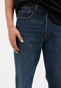 Vaqueros de denim azul oscuro con corte ajustado, que presentan costuras naranjas y un diseño clásico de cinco bolsillos. La textura parece suave y resistente.