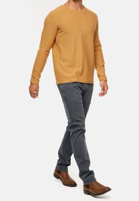 Suéter de punto de manga larga mostaza, cuello redondo, tejido texturizado, combinado con jeans gris ajustados y botas de tobillo marrones.