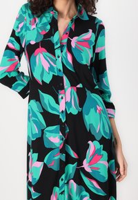 Robe à imprimé floral noire, ornée de grandes fleurs bleu sarcelle et roses. Boutons sur le devant, manches longues, coupe décontractée. Texture de tissu lisse.