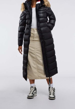 Personne portant un long manteau noir matelassé avec capuche en fourrure, jupe midi beige, chaussettes blanches et baskets noires et blanches, debout devant un fond uni.