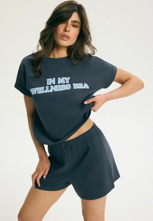 Kobieta w granatowych szortach i pasującym T-shircie z napisem "IN MY WELLNESS ERA", pozująca z jedną ręką na biodrze, a drugą lekko naciągającą koszulkę.