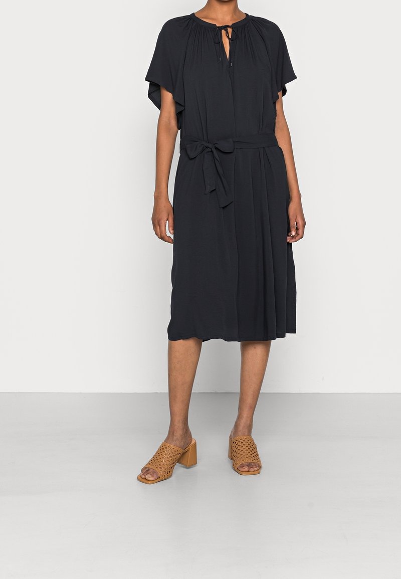 Femme portant une robe noire longueur genou avec des manches volantées, un col à nouer, une ceinture à la taille et des sandales à talons carrés tissées couleur sable.