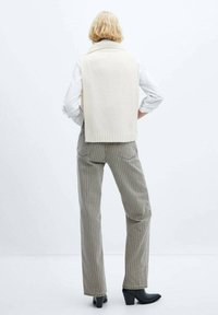 Cremesfarbenes Strickweste mit geripptem Ausschnitt, getragen über einem weißen Hemd, kombiniert mit grünen gestreiften Hosen und schwarzen Stiefeletten mit Absatz.