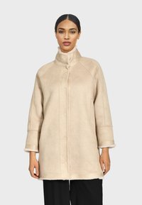 Abrigo de piel sintética beige con cuello alto, cierre delantero de botones y mangas tres cuartos con una textura suave. Diseño simple, sin patrones.