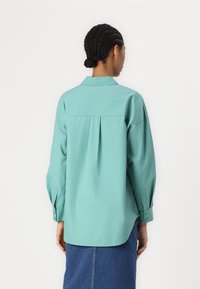 Maglietta a maniche lunghe color teal chiaro con colletto, dettaglio di piega sulla schiena e polsini abbottonati. Il tessuto sembra liscio con un design a vestibilità rilassata.