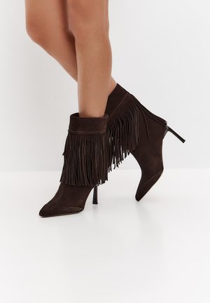 Gambe che indossano stivaletti in pelle scamosciata marrone scuro con tacco alto, con punte piegate e lunghi dettagli a frangia su uno sfondo bianco semplice.