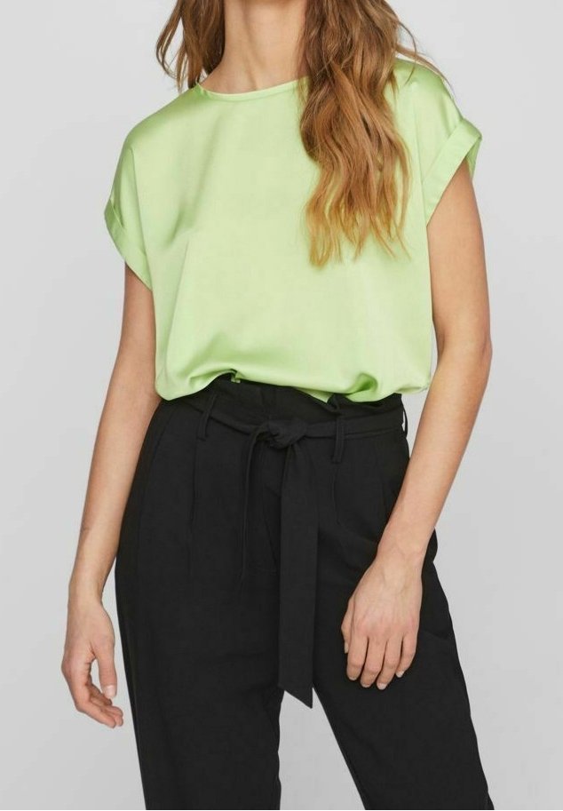 Femme portant un chemisier vert clair à manches courtes, rentré dans un pantalon noir taille haute avec une ceinture en tissu nouée devant.