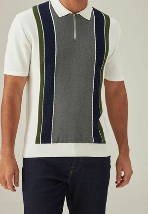 Homme portant un polo blanc à manches courtes avec des rayures verticales texturées vertes et bleu marine et une fermeture éclair à l'avant, associé à un jean bleu foncé.