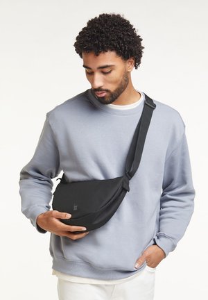 Homme portant un sweat-shirt gris clair et un pantalon blanc, tenant un sac bandoulière noir avec une sangle ajustable croisée sur sa poitrine.