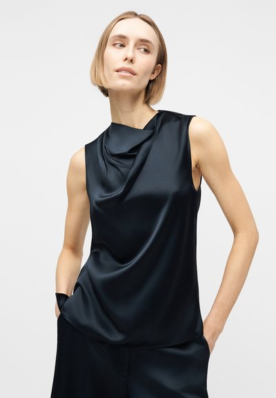 Γυναίκα με κοντά ξανθά μαλλιά που φοράει μαύρο σατέν τοπ χωρίς μανίκια με ντραπέ λαιμόκοψη, στέκεται με τα χέρια στις τσέπες του παντελονιού μπροστά σε απλό φόντο.