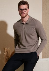 Camisa polo de manga larga en beige con cuello, dos botones y una textura suave, combinada con pantalones oscuros. Diseño moderno y ajuste entallado.