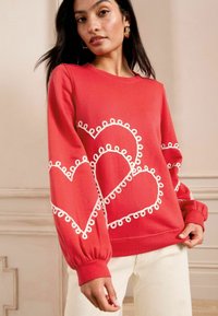 Felpa rossa con motivi a cuore bianchi e dettagli ad anello. Maniche lunghe con polsini elastici e vestibilità comoda.