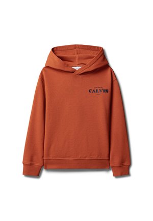 Jongen staat met handen in de zakken, draagt een roestkleurige hoodie, witte korte broek en beige sandalen tegen een grijze achtergrond.