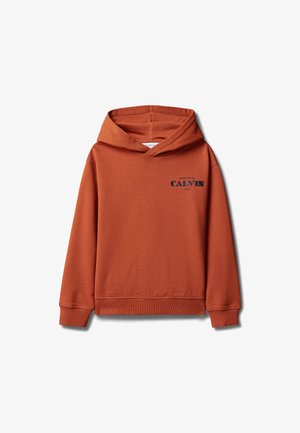 Garçon debout, les mains dans les poches, portant un sweat à capuche couleur rouille, un short blanc et des sandales beiges sur fond gris.