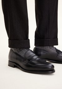 Black leather loafers with a simple design, featuring a raised band across the front. Paired with gray patterned socks and dark trousers.