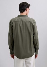 Camisa de manga larga verde oliva con cuello puntiagudo, puños abotonados y dobladillo redondeado. Textura suave, mostrada desde la parte de atrás.