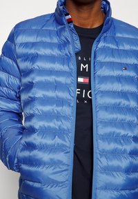 Blaue Steppjacke mit horizontalem Steppmuster, hohem Kragen und durchgehendem Reißverschluss. Verfügt über ein kleines Logo auf der linken Brustseite und eine glatte Textur.