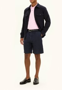 Giacca di tessuto blu navy sopra una polo rosa chiaro, abbinata a pantaloni corti blu navy. Una cintura nera e mocassini completano l'outfit, evidenziando i colori a contrasto.