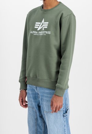 Person trägt ein olivgrünes Alpha Industries Sweatshirt mit weißem Logo und hellblauen Denim-Cargohosen vor einfarbigem Hintergrund.