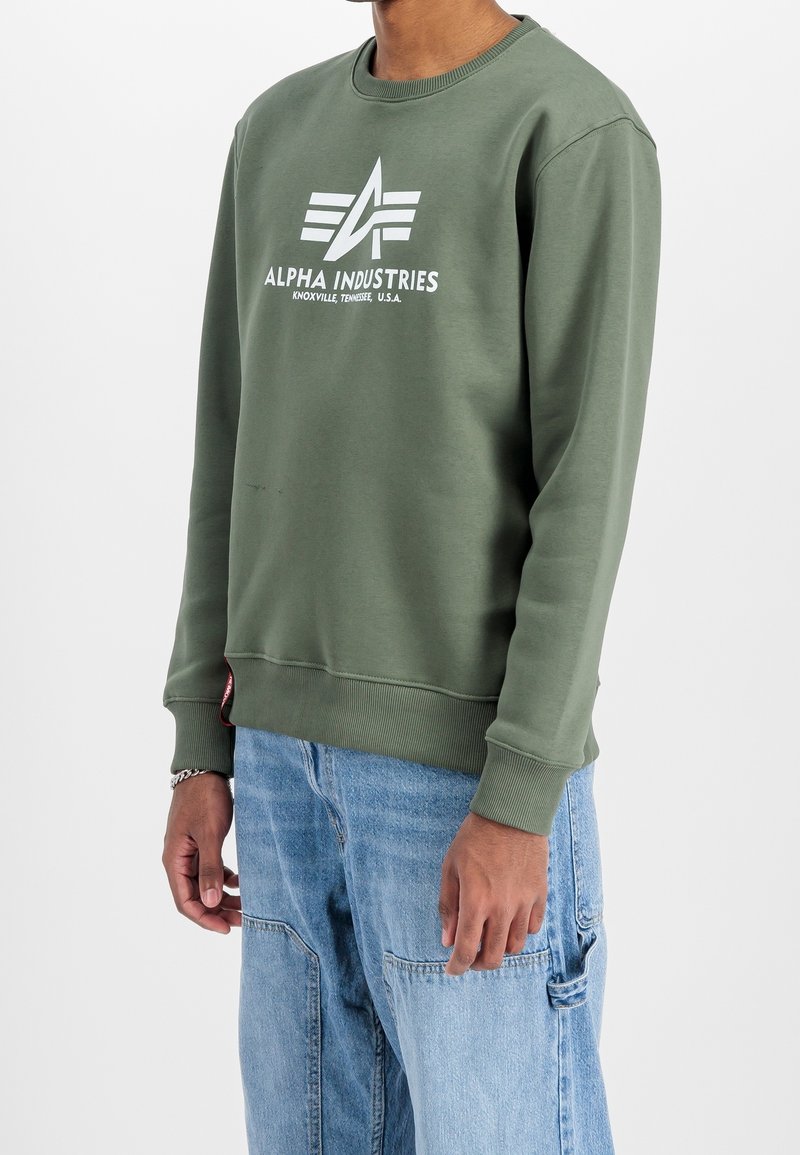 Personne portant un sweat-shirt vert olive Alpha Industries avec logo blanc et un pantalon cargo en denim bleu clair sur un fond uni.