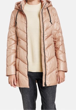 Persoon die een lichtroze doorgestikte jas met capuchon draagt over een crème trui en donkergrijze spijkerbroek, staand tegen een witte achtergrond.