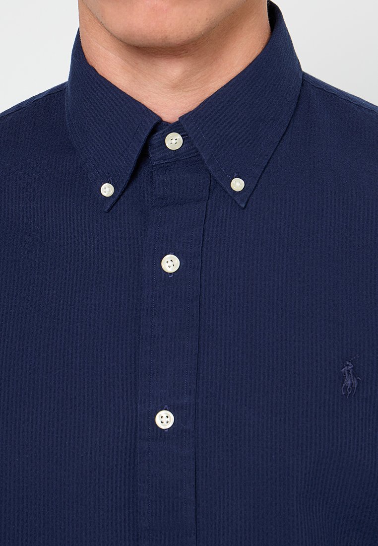 Chemise bleu marine à boutons avec un tissu texturé, un col pointu et un petit logo sur la poitrine gauche; elle présente des boutons blancs et un motif côtelé.