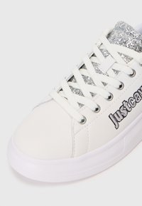 Weißer Sneaker mit glitzernden silbernen Akzenten am Kragen, ausgestattet mit weißen Schnürsenkeln, genähten Details und einer Gummisohle. Markenname sichtbar.