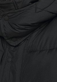 Sort polstret jakke med høj krave, trykknapper og diagonalt quiltet mønster på stoffet, der giver en glat og blød tekstur.