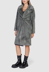 Cappotto in pelliccia sintetica grigia con colletto a punta, maniche lunghe e chiusura con un solo bottone, abbinato a stivaletti neri con zip alla caviglia.