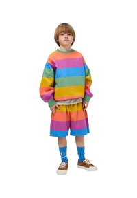 Fényes színekben csíkos pulóver és rövidnadrág szett, pink, narancssárga, zöld és kék árnyalatokkal. Középhosszú ujjú, laza szabású, kék zoknikkal és barna cipőkkel párosítva.