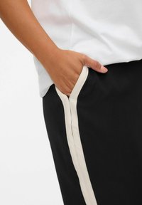 Pantalons noirs avec une rayure latérale crémeuse, présentant une main dans une poche latérale. Le tissu semble lisse avec une coupe décontractée.