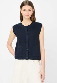 Donna che indossa un gilet lavorato a maglia blu navy senza maniche con bottoni e due tasche, e pantaloni color crema, sorridente davanti a uno sfondo chiaro e uniforme.