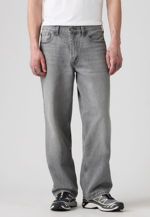 Personne portant un jean gris ample et des baskets noires et blanches, debout avec les mains détendues le long du corps sur un fond uni.
