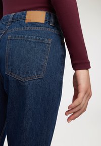 Tylny widok osoby ubranej w niebieskie dżinsy i bordową bluzkę z długim rękawem, ręka swobodnie oparta na boku, na jednolitym tle.