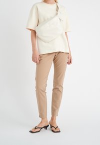 Beige bomullsmixbyxor, krämfärgad t-shirt med kort ärm och svarta flip-flops. Modellen bär också en krämfärgad axelremsväska med en elegant design.