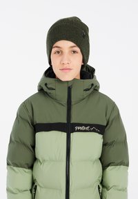 Giacca a vento verde con striscia nera, chiusura con zip e cappuccio. Berretto a coste verde scuro. Presenta un dettaglio del logo sulla giacca. Tessuto liscio.