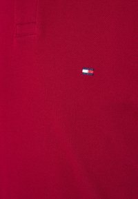 Punainen polo-paita tekstuuritassukaudesta, jossa on pieni brodeerattu logo sinisillä, valkoisilla ja punaisilla yksityiskohdilla rinnan kohdalla.