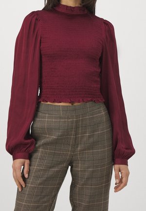 Femme portant un chemisier bordeaux ajusté à manches longues avec ourlet et poignets à volants, associé à un pantalon marron à carreaux taille haute.