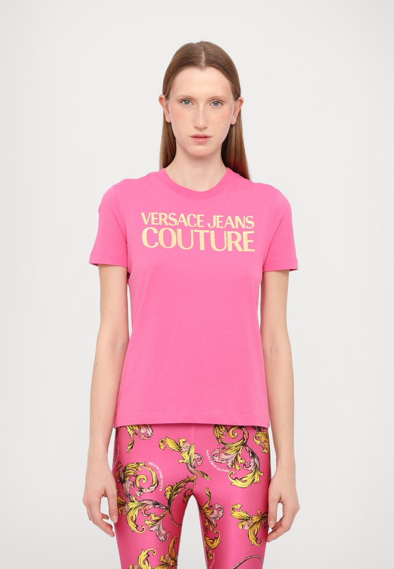 T-shirt en coton rose à manches courtes, affichant "VERSACE JEANS COUTURE" en texte jaune en gras sur la poitrine.