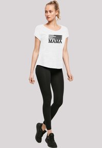 Weißes T-Shirt mit kurzen Ärmeln und schwarzem Text "STARK SCHÜTZEND GELIEBT SCHÖNE MAMA", kombiniert mit schwarzen Leggings und schwarzen Sneakern.