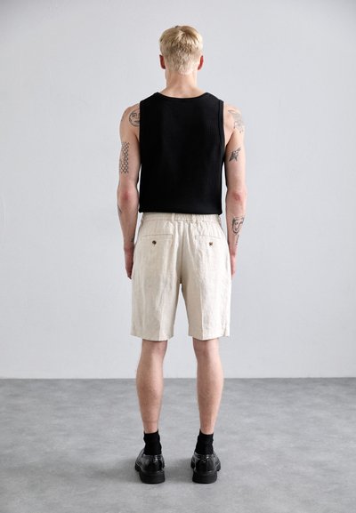 Schwarzes ärmelloses Oberteil für Herren mit gerippter Textur, beigefarbene Leinen-Shorts und schwarze Schuhe mit kontrastierenden Socken; sichtbare Rückentaschendetails.
