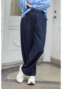 Navy wide-leg trousers with pleats and a tie waist, paired with white sneakers. Soft fabric with a smooth texture, set against a light background.